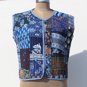 Könnte beinhalten: Eine ärmellose, gesteppte Weste mit Patchwork-Design. Die Weste weist eine Vielzahl von blauen Stoffmustern auf, darunter Blumen- und geometrische Drucke. Sie hat eine Knopfleiste vorne und eine gestreifte Bordüre. Die Weste wird auf einer Schaufensterpuppe präsentiert.