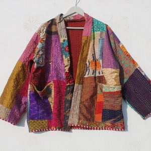 Op de afbeelding: Een kleurrijk patchwork gequilte jas met een lange, open voorkant en lange mouwen. De jas is gemaakt van verschillende stoffen in tinten paars, rood, bruin en goud.