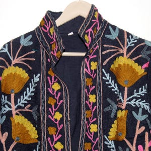 Pu&ograve; includere: Giacca blu navy scuro con ricami floreali intricati nei toni del giallo, rosa e blu. La giacca ha un collo alto ed &egrave; esposta su una gruccia di legno. Il ricamo crea uno straordinario contrasto con il tessuto scuro.