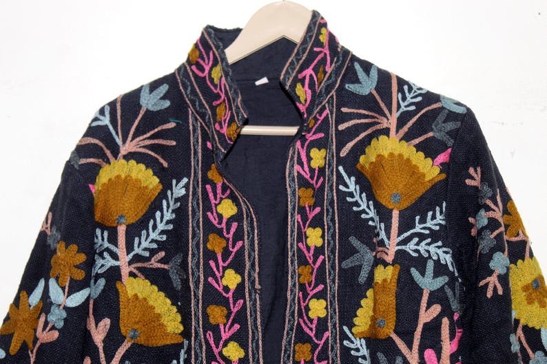 Pu&ograve; includere: Giacca blu navy scuro con ricami floreali intricati nei toni del giallo, rosa e blu. La giacca ha un collo alto ed &egrave; esposta su una gruccia di legno. Il ricamo crea uno straordinario contrasto con il tessuto scuro.