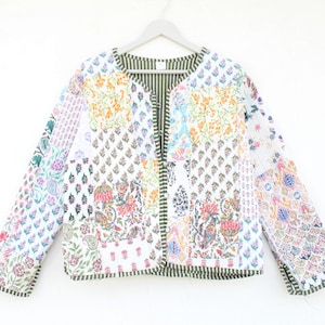 Może przedstawiać: Biała pikowana kurtka z patchworkowym wzorem, który zawiera różne kwiatowe motywy w odcieniach różowego, niebieskiego, zielonego i żółtego. Kurtka ma zieloną lamówkę wokół krawędzi i podszewkę ze wzorzystej tkaniny.