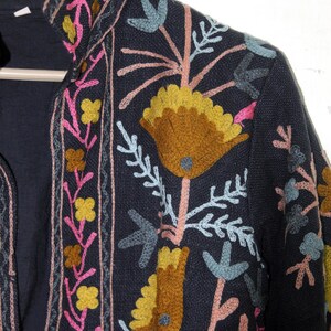 Pu&ograve; includere: Giacca blu navy scuro con ricami floreali colorati. Il ricamo presenta fiori e foglie gialle, rosa e blu. La giacca ha un collo alto ed &egrave; appesa a un'appendiabiti di legno.