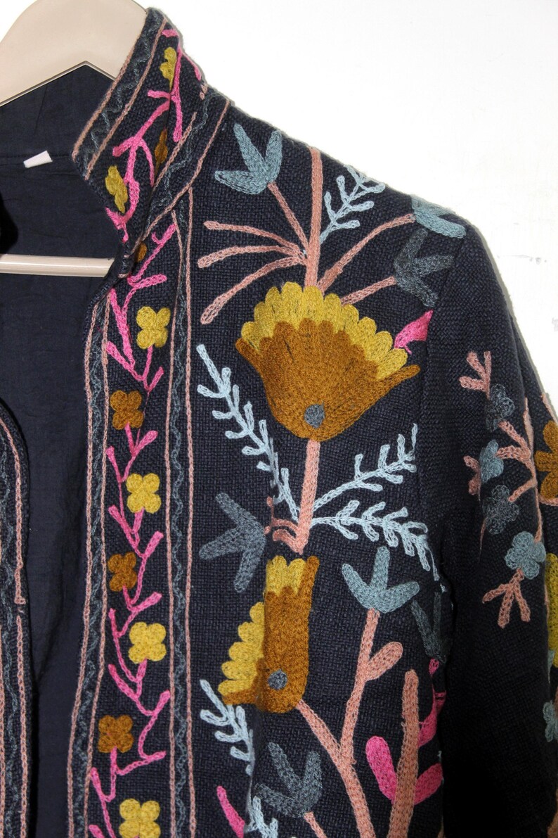 Pu&ograve; includere: Giacca blu navy scuro con ricami floreali colorati. Il ricamo presenta fiori e foglie gialle, rosa e blu. La giacca ha un collo alto ed &egrave; appesa a un'appendiabiti di legno.