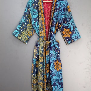 Könnte beinhalten: Ein langer, gemusterter Bademantel mit Bindegürtel, mit marineblauem Hintergrund und floralen und Paisley-Designs in Türkis- und Goldtönen. Die Innenseite des Bademantels ist einfarbig rot. Der Bademantel hängt an einem Holzbügel.