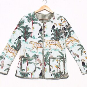 Könnte beinhalten: Eine weiße gesteppte Jacke mit einem grünen, braunen und gelben Dschungelprint mit Tukanen, Leoparden, Palmen und anderen tropischen Tieren. Die Jacke hat einen Knopfverschluss und zwei aufgesetzte Taschen.