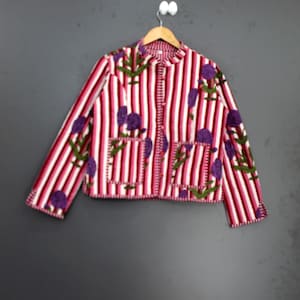 Könnte beinhalten: Gestreifte Jacke mit floralem Design. Die Jacke hat vertikale Streifen in Rosa- und Weißtönen mit lila Blumen- und grünen Blattakzenten. Sie hat zwei Vordertaschen und ist zugeknöpft.