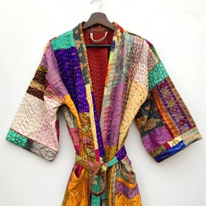 Op de afbeelding: Een kleurrijke patchwork-jas met een lange strikband. De jas is gemaakt van verschillende stoffen in tinten paars, roze, geel, groen en bruin. De stof is in een patchwork-ontwerp aan elkaar gestikt.
