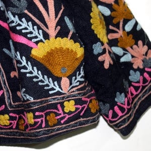 Pu&ograve; includere: Primo piano di un tessuto blu scuro con ricami floreali colorati. Il ricamo presenta fiori gialli, rosa e arancioni, con accenti azzurri e bianchi. Il design &egrave; intricato e dettagliato, con un bordo.