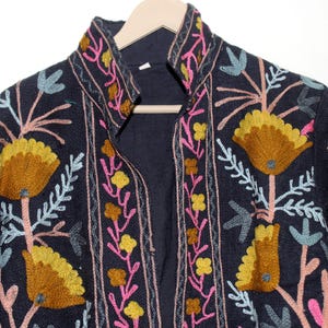 Pu&ograve; includere: Giacca blu navy scuro con ricami floreali intricati nei toni del giallo, rosa e blu. La giacca ha un collo alto ed &egrave; esposta su una gruccia di legno. Il ricamo crea uno straordinario contrasto con il tessuto scuro.