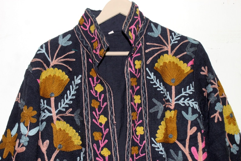 Pu&ograve; includere: Giacca blu navy scuro con ricami floreali intricati nei toni del giallo, rosa e blu. La giacca ha un collo alto ed &egrave; esposta su una gruccia di legno. Il ricamo crea uno straordinario contrasto con il tessuto scuro.