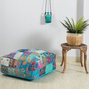 Könnte beinhalten: Ein türkisblaues Patchwork-Bodenkissen mit verschiedenen bunten Mustern und Stickereien. Das Kissen liegt auf einem hellen Holzboden vor einer weißen Wand.