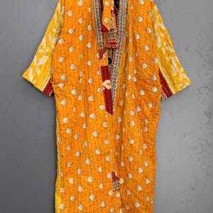 Könnte beinhalten: Ein langer, offen geschnittener Kimono in leuchtendem Gelb, verziert mit einem sich wiederholenden weißen Muster. Der Kimono hat kontrastierende rote und gemusterte Verzierungen an den Kanten und am Kragen. Er hängt an einem Holzbügel vor grauem Hintergrund.