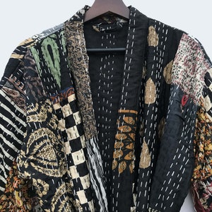Könnte beinhalten: Ein Patchwork-Kimono-Mantel mit einer Vielzahl von Mustern und Farben, darunter Schwarz, Beige, Braun und Grün. Der Mantel hängt an einem Holzbügel. Der Mantel hat weiße Nähte.