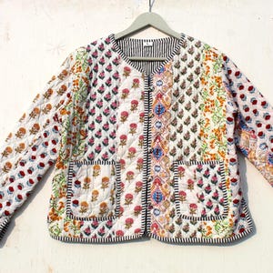 Könnte beinhalten: Eine weiße gesteppte Jacke mit Blumendruck und Knopfleiste. Die Jacke hat zwei Taschen auf der Vorderseite.