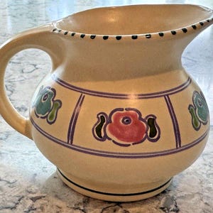 Puede incluir: Una jarra de cerámica de color crema con un diseño floral. La jarra tiene una base ancha y un cuerpo redondeado. El diseño floral presenta flores rosas, verdes y azules.