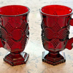Könnte beinhalten: Zwei rubinrote Glasbecher mit verzierten, geprägten Mustern und quadratischen Sockeln. Jeder Becher hat einen gebogenen Henkel und ein detailliertes Design am Körper. Die Becher werden auf einer hellen Oberfläche präsentiert.
