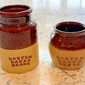 Op de afbeelding: Set van twee vintage keramische potten. De potten zijn bruin en beige met de tekst "BOSTON BAKED BEANS" op de voorkant. Eén pot is hoog en cilindrisch, de andere is een kleine, ronde pot.