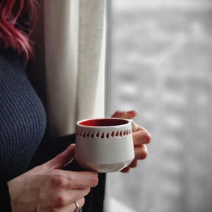 Op de afbeelding: Een witte keramische mok met een rode rand en een decoratief patroon van rode driehoeken rond de bovenkant. De mok wordt vastgehouden door een hand.