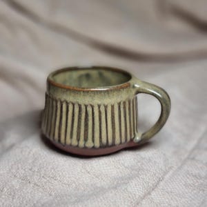 May include: A small, handmade ceramic mug with a rounded handle. The mug features vertical, textured lines in a light green and brown color scheme. The base is a darker brown, and the interior has a green hue.