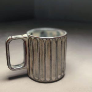 Puede incluir: Una taza de cerámica con un diseño de costillas verticales en tonos grises y marrones. La taza tiene un asa rectangular y un borde ligeramente ensanchado. El interior de la taza tiene un tono marrón rojizo.