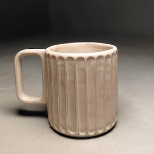 Può includere: Una tazza in ceramica beige con dettagli a coste verticali. La tazza ha un manico quadrato e una forma cilindrica. Il colore neutro e il design semplice la rendono adatta a varie bevande e all'arredamento della cucina.