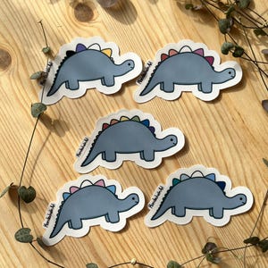 Könnte beinhalten: Fünf Aufkleber mit einem Cartoon-Dinosaurier mit grauem Körper und farbenfrohem Rückgrat. Die Aufkleber befinden sich auf einer Holzoberfläche mit Grünzeug. Jeder Dinosaurier hat ein anderes Regenbogenfarbschema auf dem Rücken.