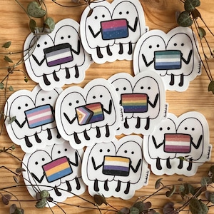 Könnte beinhalten: Eine Sammlung von gestanzten Vinyl-Aufklebern, die Paare von weißen, geisterähnlichen Figuren zeigen, die verschiedene Pride-Flaggen halten. Jeder Aufkleber zeigt ein einzigartiges Flaggendesign mit verschiedenen Farben, Streifen und Verläufen.