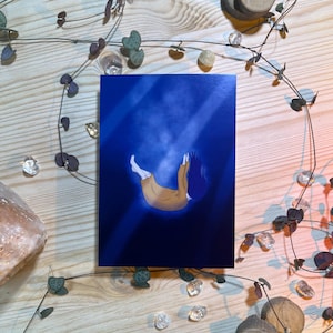 Könnte beinhalten: Ein Kunstdruck, der eine Person zeigt, die in einen dunkelblauen Raum mit Lichtstrahlen fällt. Die Person trägt ein beiges Outfit mit blauen Haaren. Der Druck ist von dekorativen Elementen umgeben, darunter klare Kristalle, Holzscheiben und eine Ranke.