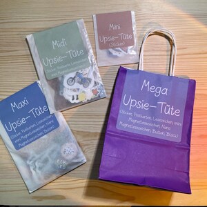 Könnte beinhalten: Draufsicht auf vier Pakete auf einer Holzoberfläche. Die Pakete sind mit "Maxi", "Midi", "Mini" und "Mega Upsie-Tüte" beschriftet. Das "Mega"-Paket ist eine lila Papiertüte mit einem weißen Etikett. Die anderen Pakete sind transparent mit farbigen Etiketten.