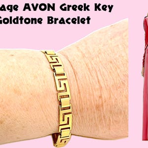 Op de afbeelding: Vintage goudkleurige armband met een Grieks sleutelpatroon. De armband wordt om een pols getoond. De afbeelding bevat de tekst "Vintage AVON Greek Key Goldtone Bracelet". Een vrouw in een roze jurk staat op de achtergrond.