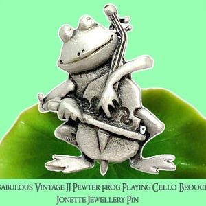 May include: A silver-coloured pewter frog brooch playing a cello, set on a green leaf against a light green background. The brooch has the text "FABULOUS VINTAGE JJ PEWTER FROG PLAYING CELLO BROOCH" and "JONETTE JEWELLERY PIN".