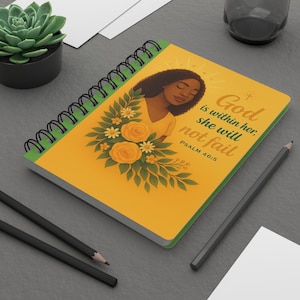 May include: Spiral-bound notebook with a yellow cover featuring an illustration of a woman with flowers and the text "God is within her, she will not fail" from Psalm 46:5. The notebook has a green spine and black spiral binding.