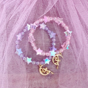 May include: Two iridescent star-shaped bracelets in a pink and purple colour scheme. The bracelets are layered on top of each other and have a charm attached to the bottom.