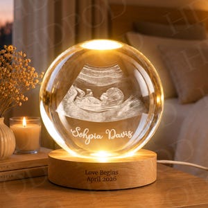 May include: A spherical glass ornament featuring a 3D laser-etched ultrasound image of a fetus inside. The glass sphere is engraved with the name Sophia Davis and sits on a circular wooden base inscribed with the text Love Begins April 2026.