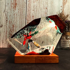 May include: A colorful robot figurine encased in a clear, geometric resin block. The robot is red, black, and yellow, with a green weapon. The resin block sits on a wooden base, creating a unique display piece.