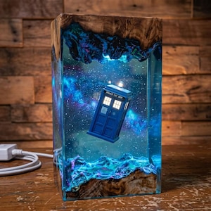 May include: A rectangular resin and wood lamp featuring a blue police box miniature suspended in a clear, glowing blue space-themed interior with starry nebula patterns. The top and bottom edges are crafted from natural wood with a polished finish.