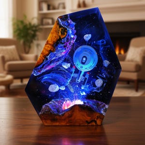 Puede incluir: Lámpara hexagonal de resina que representa una escena espacial con una nave espacial azul, planetas y asteroides. La lámpara tiene una base de madera y está iluminada con luz azul, creando un efecto de galaxia. La lámpara está sobre una mesa de madera.