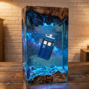 May include: A rectangular lamp featuring a blue police box suspended within a clear resin block. The resin contains a swirling nebula of blue and purple hues with white stars. The top and bottom are encased in natural wood with organic edges.