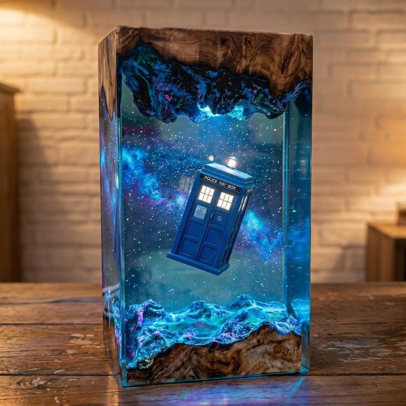 May include: A rectangular lamp featuring a blue police box suspended within a clear resin block. The resin contains a swirling nebula of blue and purple hues with white stars. The top and bottom are encased in natural wood with organic edges.