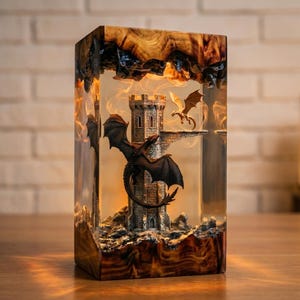 Pu&ograve; includere: Una lampada rettangolare in resina con base e parte superiore in legno naturale. All'interno della resina trasparente, una torre color pietra poggia su una base rocciosa. Un grande drago nero avvolge la torre, mentre un drago dorato vola.