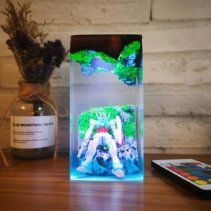 May include: A rectangular resin art piece with a white and red robot figure set within a miniature green landscape. The piece is illuminated with blue light, creating a vibrant display. A remote control is placed nearby.