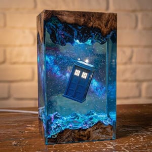 May include: A rectangular lamp featuring a blue police box model suspended within a clear resin block. The resin contains a swirling blue and purple nebula design with stars. The top and bottom edges are framed by natural wood with rough edges.