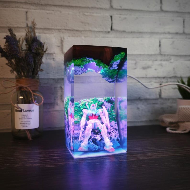 May include: A rectangular resin lamp with a white and red robot figure set in a green and blue landscape. The lamp has a dark wood top and emits a soft, purple glow. The lamp is on a dark surface.