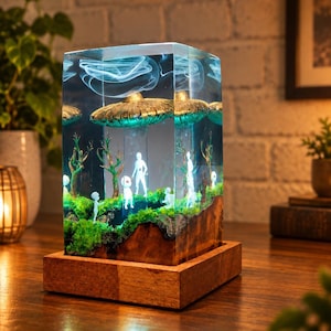 May include: A rectangular resin lamp featuring a diorama of a golden UFO hovering over a green mossy landscape with small white alien figures and bare trees. The scene is encased in clear resin with a swirling white effect and a wooden base.