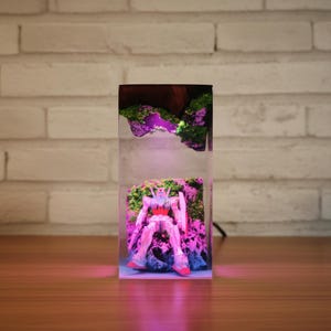 May include: A clear resin block contains a detailed robot figure set amongst miniature rock formations and moss. The scene is lit with a pink and purple glow, creating a unique decorative piece.