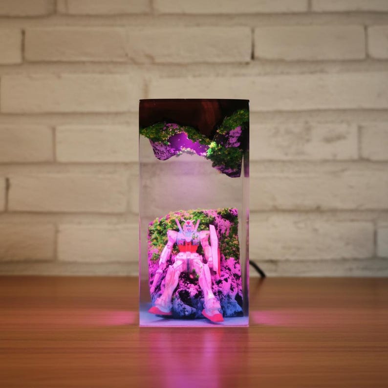 May include: A clear resin block contains a detailed robot figure set amongst miniature rock formations and moss. The scene is lit with a pink and purple glow, creating a unique decorative piece.