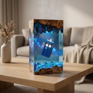May include: A rectangular lamp made of natural wood and blue resin, featuring a miniature blue police box floating in a cosmic scene with stars and nebulae. The resin interior glows with light, illuminating the detailed blue box structure.