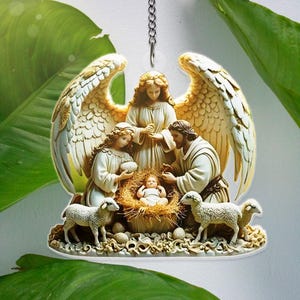 May include: A religious ornament depicting the Nativity scene. The ornament features an angel with outstretched wings, figures of Mary, Joseph, and baby Jesus in a manger, and several sheep. The figures are in cream and gold tones.