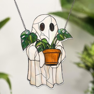 May include: A stained glass ghost holding a potted plant with green leaves. The ghost is white with black eyes and a chain hanging from the top.