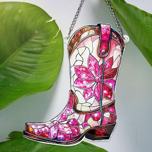 May include: A pink and white stained glass cowboy boot with floral designs. The boot is hanging from a silver chain.